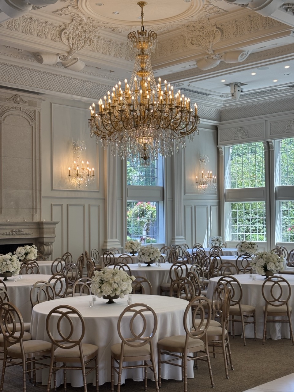Elegant ballroom at Florentine Gardens wedding venue in River Vale, New Jersey, featuring crystal chandeliers, round reception tables with white linens, white floral centerpieces, and floor-to-ceiling windows overlooking lush gardens.